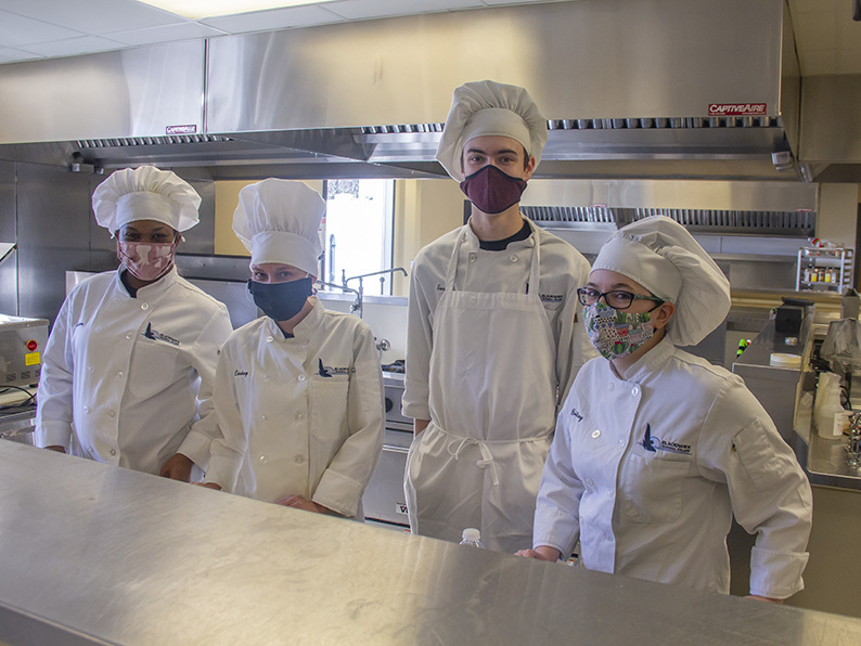 Four uniformed culinary arts students stand in a row in the BTC culinary kitchen
