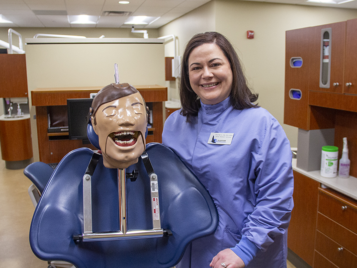 Kristen, a woman with brown shoulder length hair wearing blus medical scrubs poses with a dental mannekin