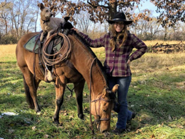 Brianna stands next to her horse, and her dog is sitting in the saddle