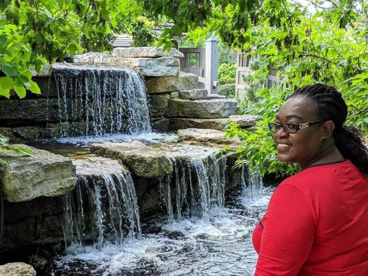 Cynthia, a black woman wearing a red short-sleeved top stands in front of a waterfall