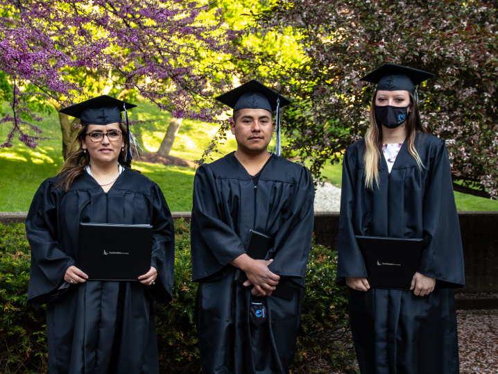 three students stand in the courtyard in their caps and gowns, holding their diplomas