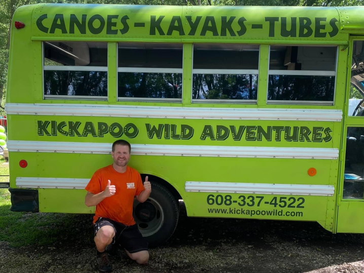 Shaun, in an orange shirt and black shorts, kneels in front of a green shuttle bus giving a thumbs up