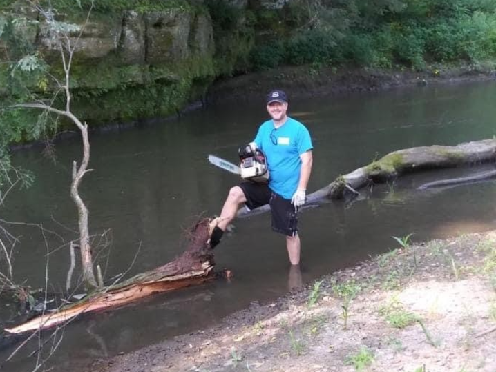 Shaun beside the river in an aqua t-shirt and black shorts holding a chainsaw