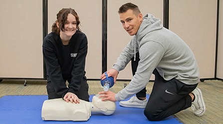 Two students performing CPR on a dummy looking at the camera
