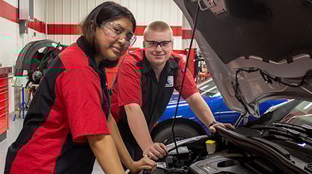 Two students smiling at camera standing over the open hood of a car