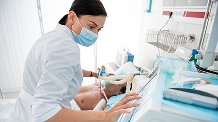 Female medical worker in protective mask touching monitor of mechanical ventilator.