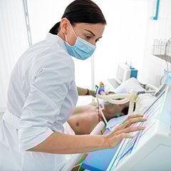 Female medical worker in protective mask touching monitor of mechanical ventilator.