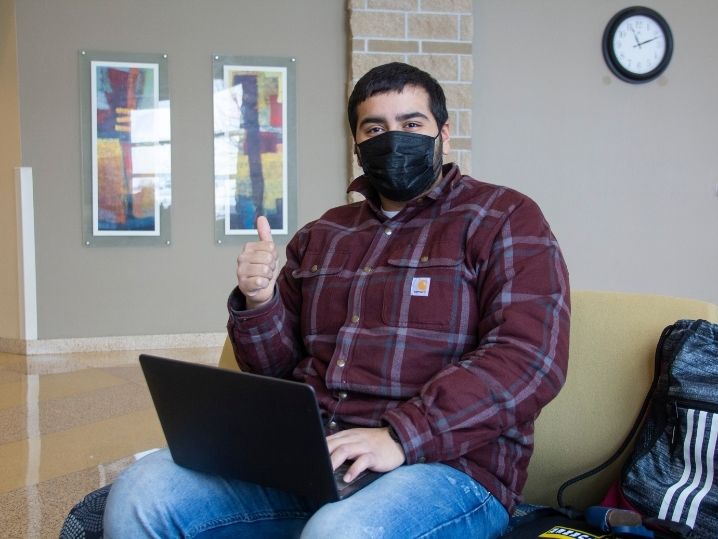 male student masked in lobby using laptop