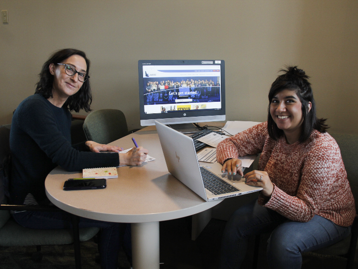 two students at a desk, one using a laptop